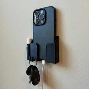 May include: A black wall-mounted phone holder with a phone, keys, and a lip balm. The phone is in a black case and is connected to a white charging cable. The holder has slots for the phone, keys, and lip balm.