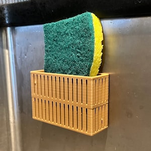 May include: A brown plastic sponge holder with a green and yellow sponge inside. The holder is attached to a stainless steel surface.