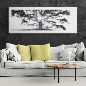 Peut inclure: Un salon moderne avec un canapé gris clair orné de coussins blancs et jaunes. Une table basse ronde en bois avec des pieds noirs est placée devant le canapé. Au-dessus, une grande photographie encadrée en noir et blanc d'un arbre.