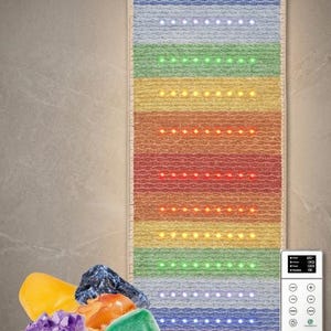 May include: A rectangular wellness mat featuring horizontal bands of textured fabric in rainbow colors, embedded with rows of small LED lights. A digital controller with buttons and a screen is attached. A collection of various polished gemstones sits nearby.