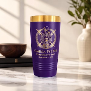 May include: A purple tumbler with a gold rim featuring the gold Omega Psi Phi Fraternity, Inc. crest and the text November 17, 1911. The cylindrical vessel has horizontal ridges near the base and a smooth finish on the upper portion of the body.