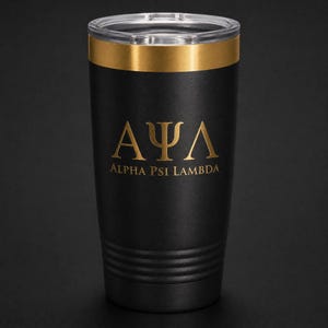 May include: A matte black stainless steel tumbler featuring a gold-colored rim and a clear lid. The front displays the gold Greek letters Alpha Psi Lambda above the text Alpha Psi Lambda in a matching gold font, with horizontal ridges at the base.