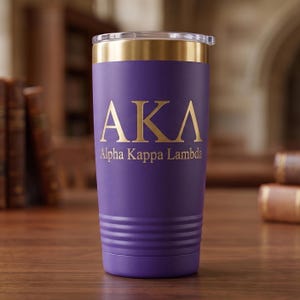 May include: A purple stainless steel tumbler with a gold-colored rim and a clear lid. The front features the gold-colored letters AKA and the text Alpha Kappa Lambda printed below. The base of the tumbler has a series of horizontal ridges.