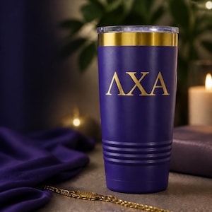 May include: A purple stainless steel tumbler featuring a gold-colored rim and the Greek letters AXA printed in gold on the front. The cylindrical vessel has a series of horizontal ridges near the base and a clear plastic lid on top.