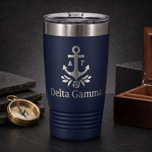 May include: A navy blue stainless steel tumbler with a clear lid, featuring a laser-engraved silver anchor emblem, Greek letters Delta Gamma, and the text Delta Gamma. The cylindrical cup has horizontal ridges near the base.