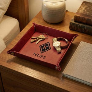 Puede incluir: Una bandeja de cuero burdeos con un diseño en forma de diamante que contiene la letra "K" y la palabra "NUPE". La bandeja contiene llaves, un reloj dorado y una pulsera marrón. La bandeja está sobre una mesita de noche de madera.