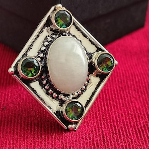 May include: Silver-toned ring with a central oval, off-white stone, surrounded by four round, green gemstones. The ring has a diamond-shaped frame with a beaded border, set against a red fabric background.