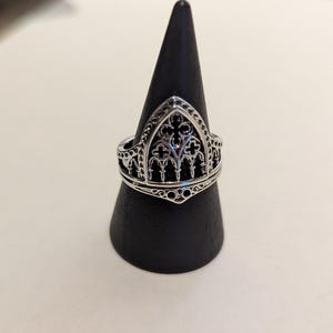May include: A silver-colored ring with a gothic cathedral design. The ring features intricate detailing, including arched windows and crosses. The ring is displayed on a black, cone-shaped stand.