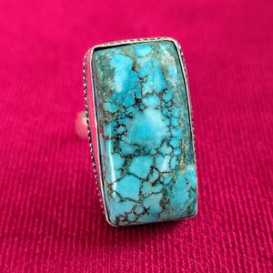 May include: A rectangular turquoise ring with a silver band. The turquoise stone has a vibrant blue color with brown veining. The ring is set against a pink background.