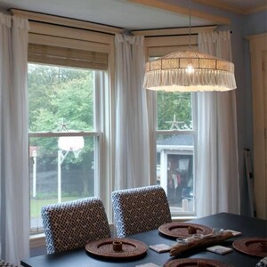 May include: A woven pendant light with a fringed bottom hangs above a dining table. The light fixture is suspended from the ceiling and illuminates the dining area. White curtains frame the windows, and patterned chairs surround the table.