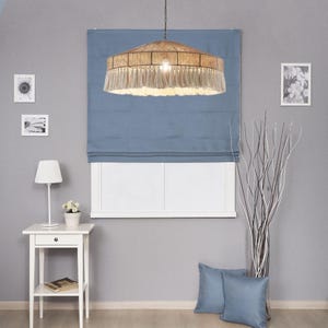May include: A woven pendant light with a fringed bottom hangs above a blue window shade. The room features a white side table with a lamp and a small vase of flowers. Two blue throw pillows rest on the floor.