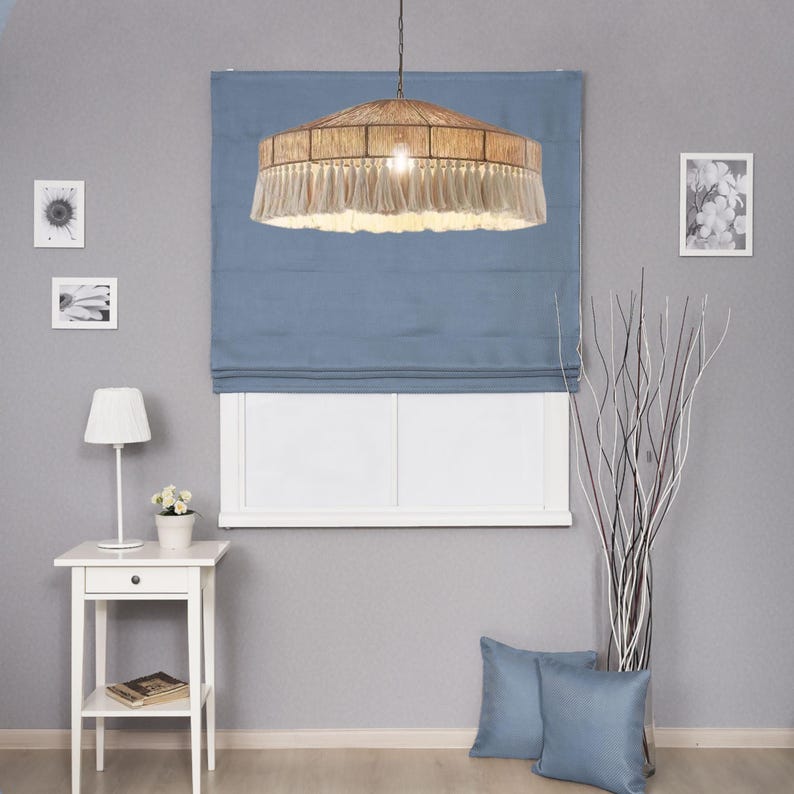 May include: A woven pendant light with a fringed bottom hangs above a blue window shade. The room features a white side table with a lamp and a small vase of flowers. Two blue throw pillows rest on the floor.