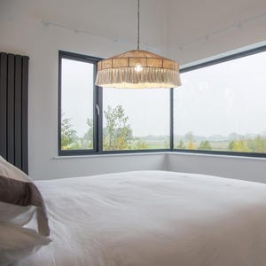 May include: A beige and cream-colored pendant light fixture with a woven shade and fringed trim hangs from the ceiling. The light is centered in a room with a large window overlooking a landscape.