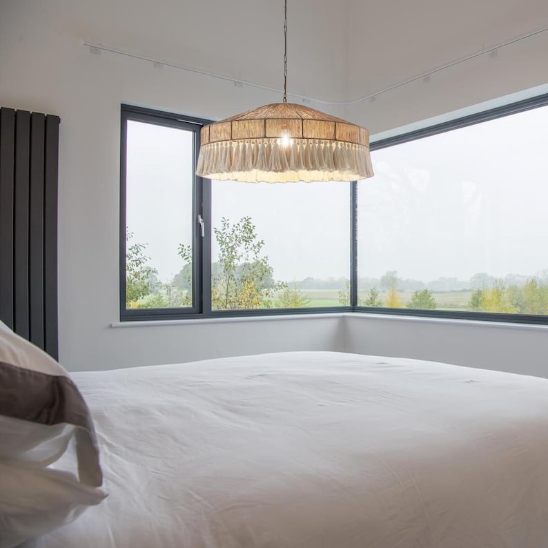 May include: A beige and cream-colored pendant light fixture with a woven shade and fringed trim hangs from the ceiling. The light is centered in a room with a large window overlooking a landscape.