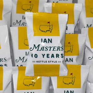 May include: A white snack bag with yellow vertical stripes featuring a graphic of the United States with a golf flag. The text reads IAN MASTERS 40 YEARS KETTLE STYLE in dark green lettering. The bag is surrounded by identical snack bags.