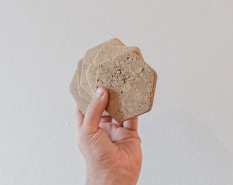 May include: A set of six beige hexagon-shaped coasters made of natural stone. The coasters are stacked on top of each other.