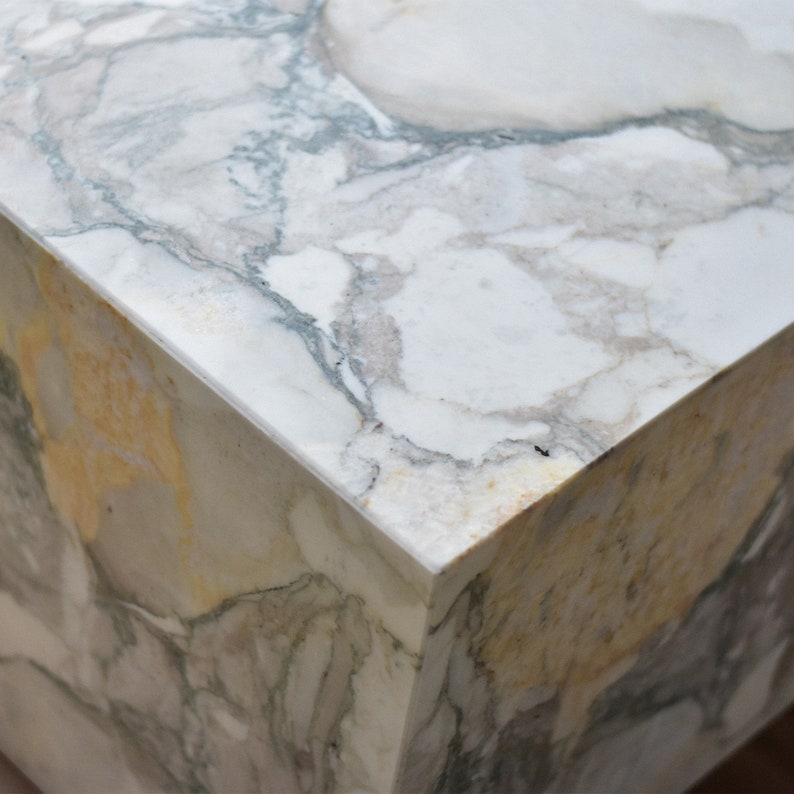 May include: Close-up of a white marble table with grey and yellow veining. The table has a square shape and a smooth, polished surface.