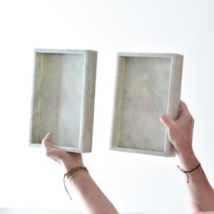 May include: Two white marble rectangular trays with slightly rounded edges. The trays are held up by two hands.