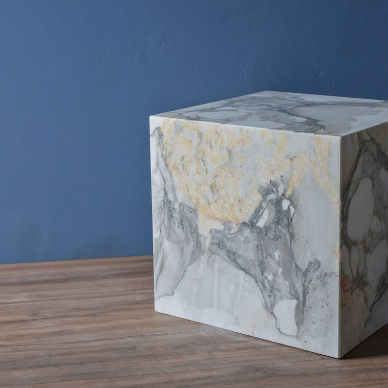 May include: A white marble cube with grey and yellow veining. The cube is sitting on a wooden surface.