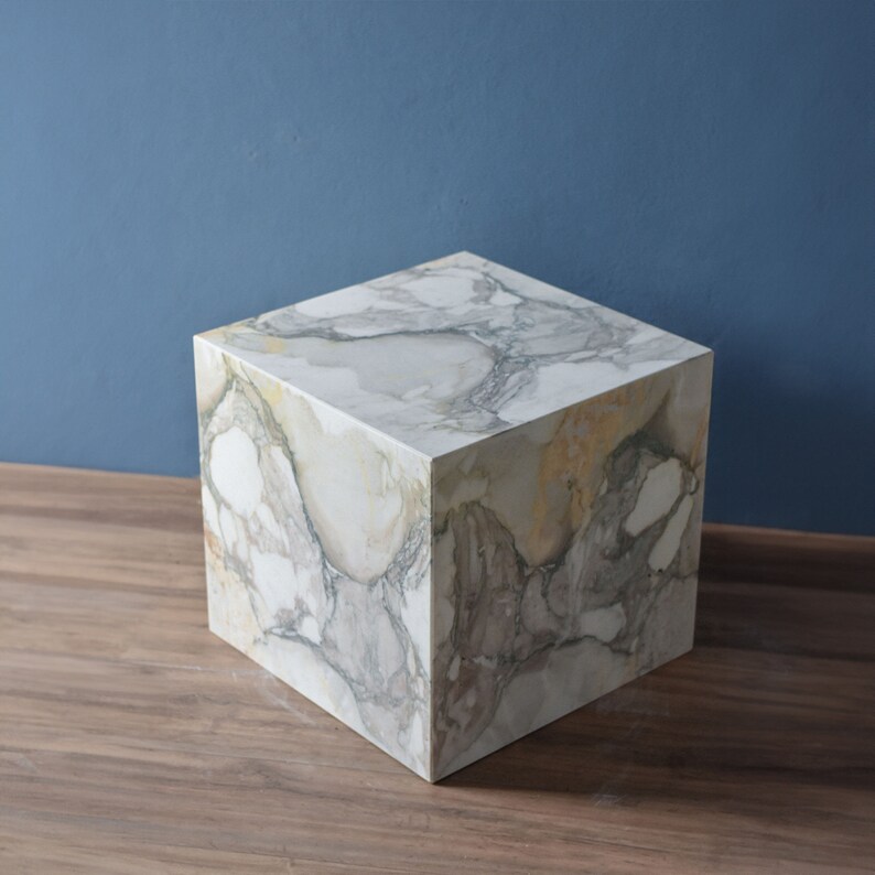 May include: A white marble cube with grey and yellow veining. The cube is sitting on a wooden surface.