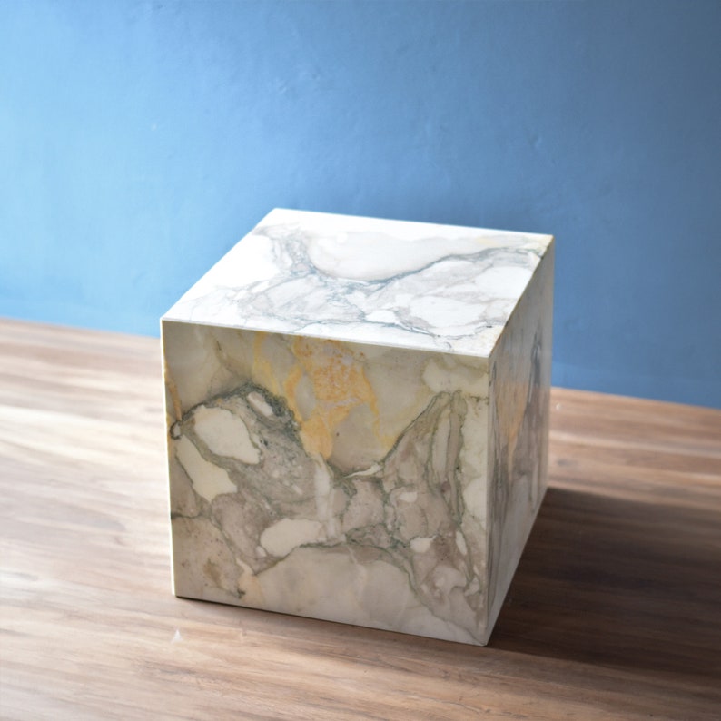 May include: A white marble cube-shaped side table with grey and gold veining. The table is sitting on a wooden floor.
