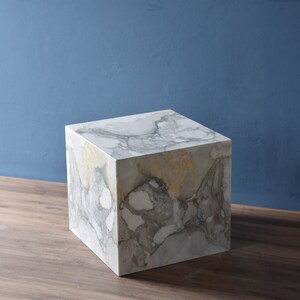 May include: A white marble cube with grey and yellow veining. The cube is sitting on a wooden surface.
