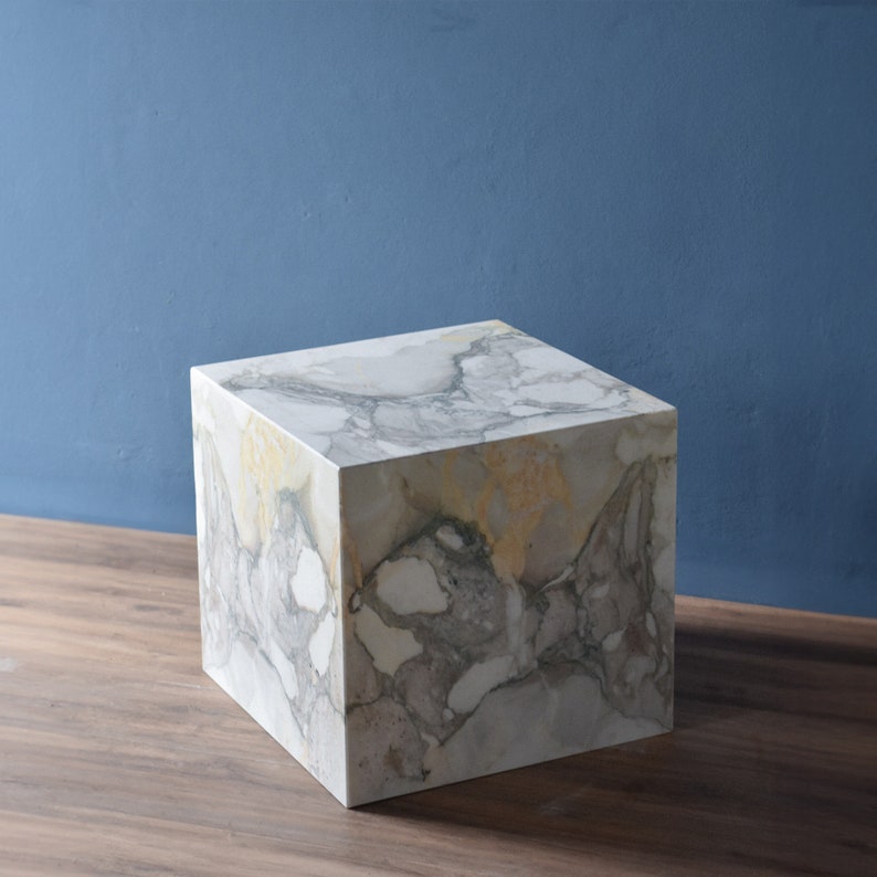 May include: A white marble cube with grey and yellow veining. The cube is sitting on a wooden surface.