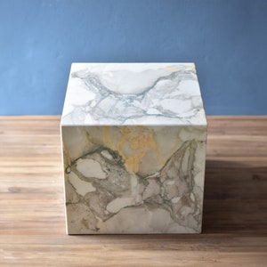 May include: A white marble cube-shaped side table with grey and gold veining. The table is sitting on a wooden surface.