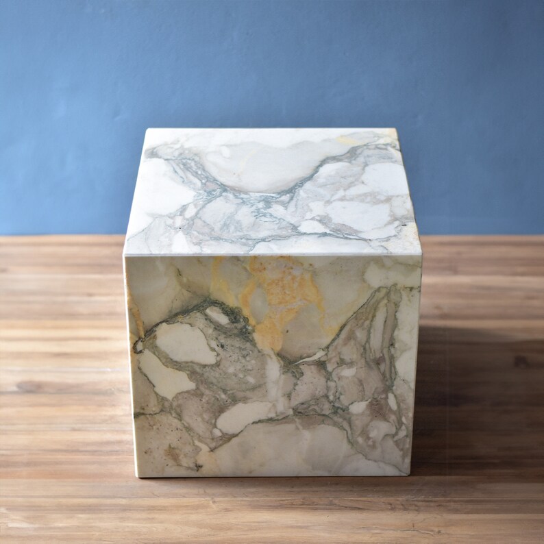 May include: A white marble cube-shaped side table with grey and gold veining. The table is sitting on a wooden surface.