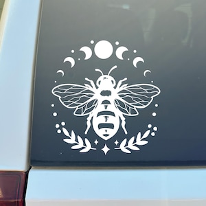 May include: White decal featuring a bee with detailed wings, surrounded by a crescent moon and moon phase design. The bee is centered within a circle of leaves and stars. The design is on a dark background.