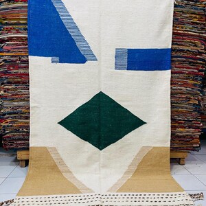 May include: A rectangular, off-white rug with geometric designs in blue, green, and tan. The rug features a large green diamond shape, blue angular shapes, and tan triangles. The bottom edge has a fringe.