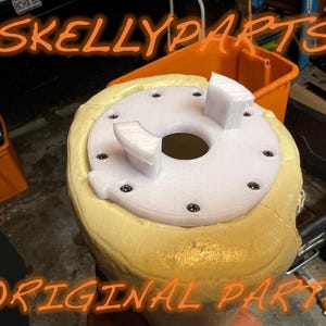 May include: A close-up of a white and yellow object with the text "SKELLYPARTS ORIGINAL PARTS" in orange. The object has a white circular top with black screws and a central hole. The object is surrounded by a yellow foam-like material.