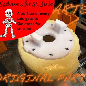 May include: A cylindrical object with a textured tan exterior and a white circular top plate secured by six screws. The plate features two raised white brackets and a central circular opening. Text on the image reads Skeletons for St. Jude.