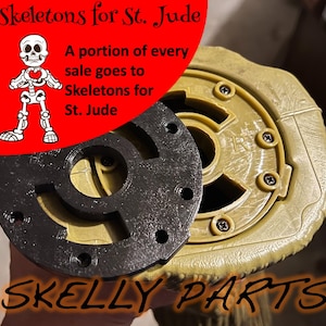 May include: A black circular plastic component with multiple screw holes positioned over a tan, textured, circular base with visible screws. The image includes a red graphic with a skeleton illustration and the text Skeletons for St. Jude and SKELLY PARTS.
