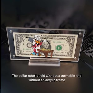 May include: A framed one-dollar bill with a cartoon character pointing at a table of gold coins. The bill is displayed in a clear acrylic frame with a clear stand. The text on the bill is visible.