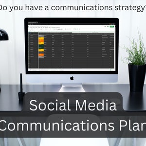 May include: A computer screen displaying a spreadsheet with the text "Social Media Communications Plan" on a black desk with a lamp, a vase with white flowers, and a potted plant.