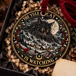 Puede incluir: Un adorno redondo de cerámica con un gato negro de ojos amarillos brillantes sobre un pueblo nevado. El texto dice THE YULE CAT IS WATCHING. El diseño incluye piñas, bayas rojas, ovillos de lana y una cinta de terciopelo rojo.