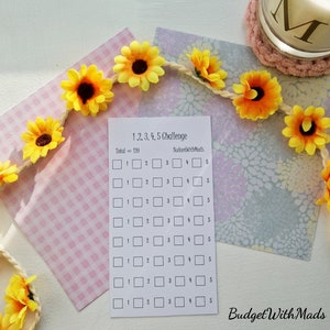 May include: A white paper with a "1,2,3,4,5 Challenge" checklist and the text "BudgetWithMads". A garland of yellow sunflowers and a pink gingham paper are also in the image. A candle with the letter "M" is in the background.