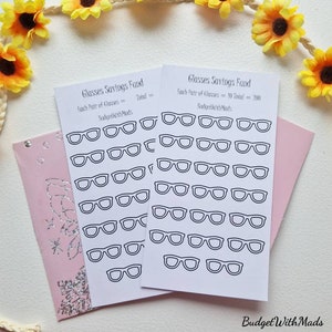 May include: Two white cards with black line drawings of eyeglasses are displayed, labeled "Glasses Savings Fund." Each card has a pink envelope. A decorative garland of yellow sunflowers and white rope surrounds the cards.