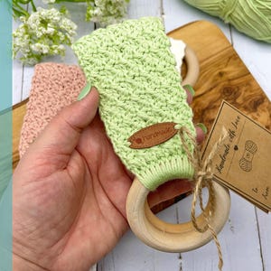 Puede incluir: Un mordedor de bebé de ganchillo verde claro con un anillo de madera. El mordedor tiene una pequeña etiqueta de cuero marrón que dice "handmade". Una pequeña etiqueta con las palabras "handmade with love" está adjunta con hilo. La palabra "PATTERN" está escrita verticalmente.