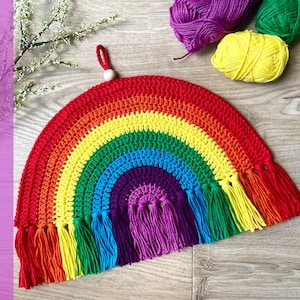 Op de afbeelding: Een kleurrijk regenboog wandkleed gehäkeld met een houten kraal aan de bovenkant en kwastjes aan de onderkant. De regenboog is gemaakt van rood, oranje, geel, groen, blauw, indigo en violet garen.