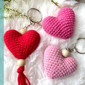 May include: Three crocheted heart-shaped keychains, one red, one pink, and one light pink. The red keychain has a wooden bead and a red tassel. The pink keychains have wooden beads and silver key rings.