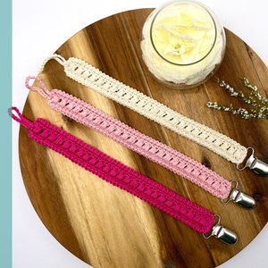 May include: Three crocheted pacifier clips in pink, light pink, and white with silver clips. The clips are laying on a wooden surface.