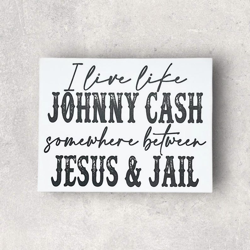 May include: White rectangular sign with black text that reads "I live like JOHNNY CASH somewhere between JESUS & JAIL". The text is in a mix of cursive and bold fonts.