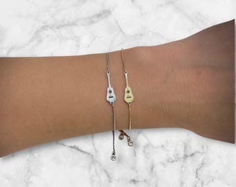 Könnte beinhalten: Zwei zarte Gitarrenarmbänder, eines in Silber und eines in Gold, werden gezeigt. Die Armbänder haben eine feine Kette und kleine, baumelnde Anhänger. Der Hintergrund ist eine marmormusterte Oberfläche, die das Design des Schmucks hervorhebt.