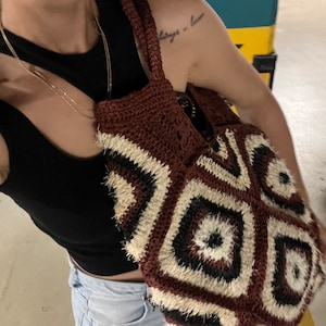 May include: A handmade tote bag with a geometric pattern. The bag features a repeating diamond design in shades of brown, black, and cream. The bag has a textured, fringed appearance and a woven handle. The bag is carried over the shoulder.