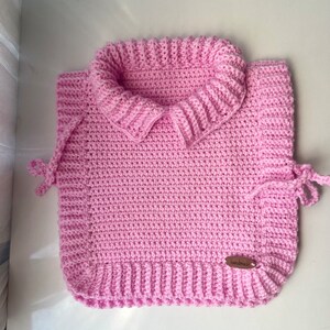 Może przedstawiać: Różowa kamizelka wykonana na szydełku z kołnierzem i sznurkami. Kamizelka ma brązową metkę z napisem "ręcznie robione".