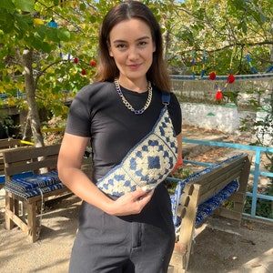 May include: A cream and navy blue crochet crossbody bag with a geometric pattern. The bag has a long strap and is held by a person wearing a black top and trousers. A gold chain necklace with purple accents is also visible.