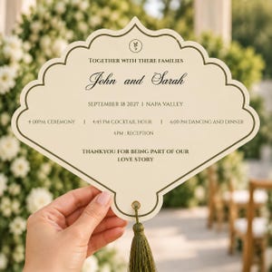 May include: A cream-colored, scalloped-edge wedding program fan with dark brown text. It features the names John and Sarah, the date September 18 2027, location Napa Valley, event schedule, and a green tassel attached through a gold metal eyelet.