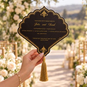 May include: A black fan-shaped wedding invitation card with gold foil lettering and decorative borders. The text reads: Together with their families, John and Sarah, September 18 2027, Napa Valley, 4:30 PM Ceremony, 4:45 PM Cocktail Hour, 6:00 PM Dancing and Dinner, 6PM Reception, Thank you for being part of our love story. A gold tassel hangs from the bottom.